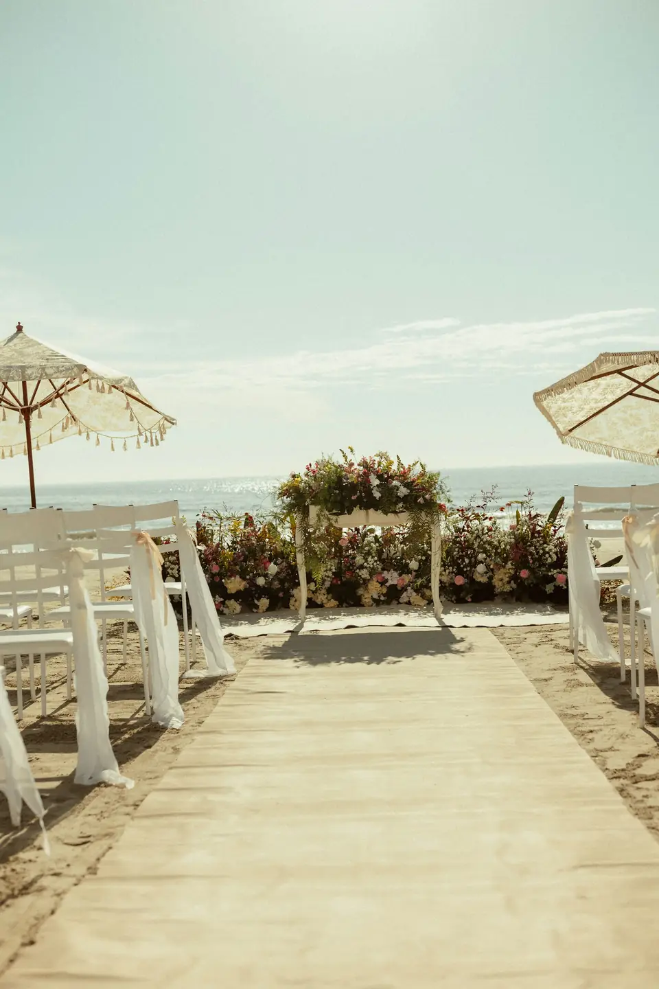 Pasillo de ceremonia en la playa con sillas blancas y arco de flores frente al mar